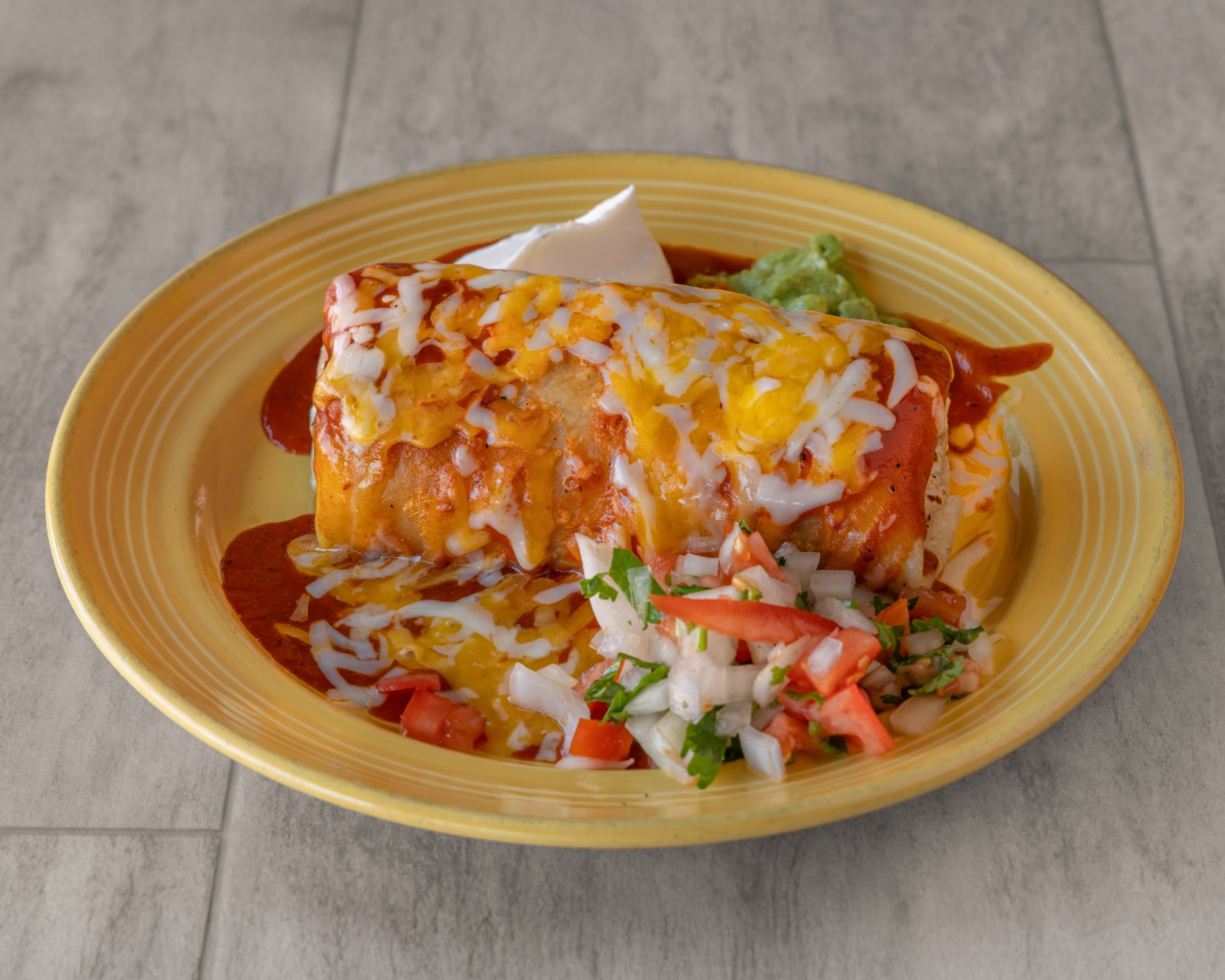 A burrito covered in red sauce and melted cheese with pico de gallo, sour cream and guacamole on a yellow plate