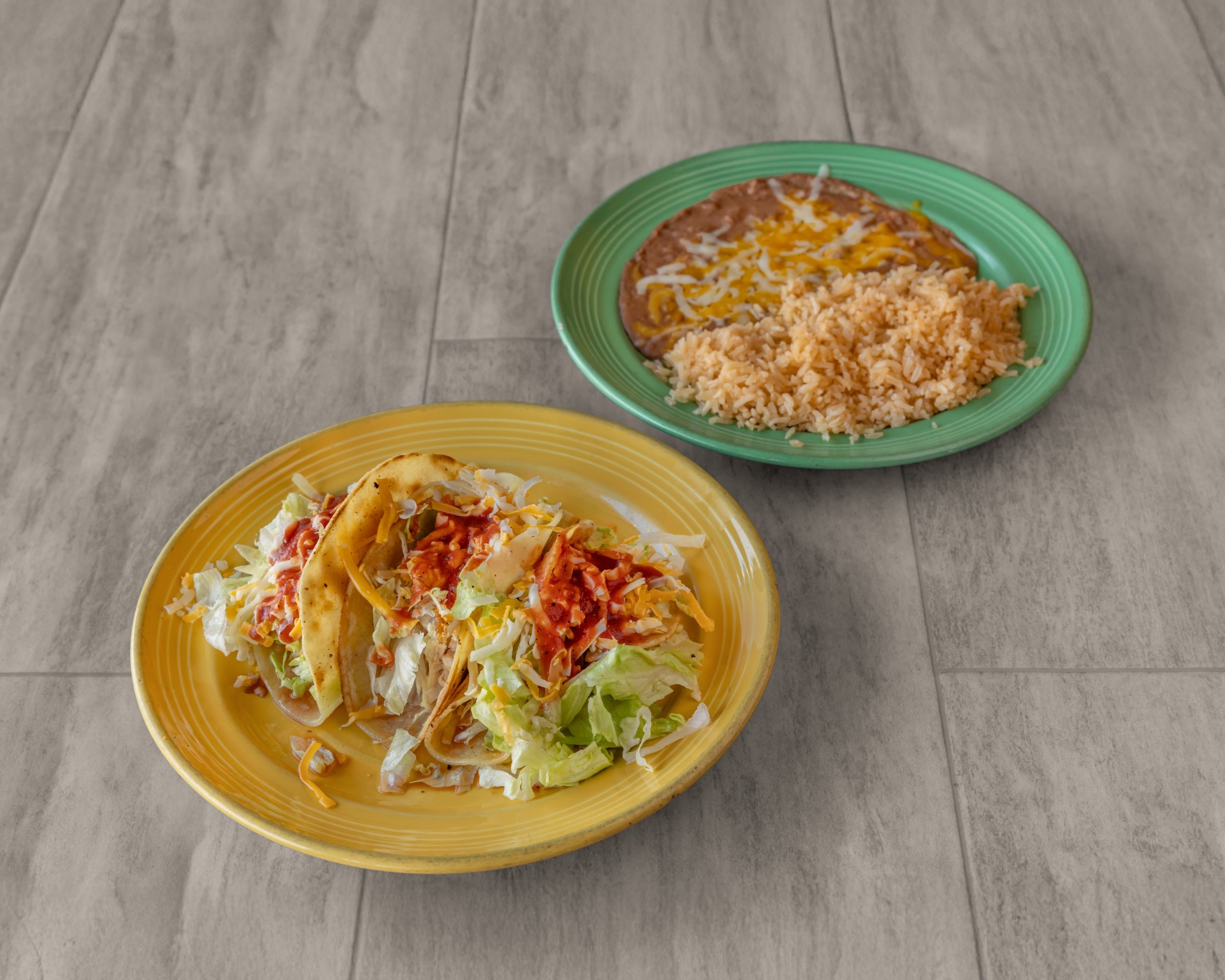 Two crispy tacos with lettuce, cheese and red sauce on a yellow plate, with rice and beans on a green plate