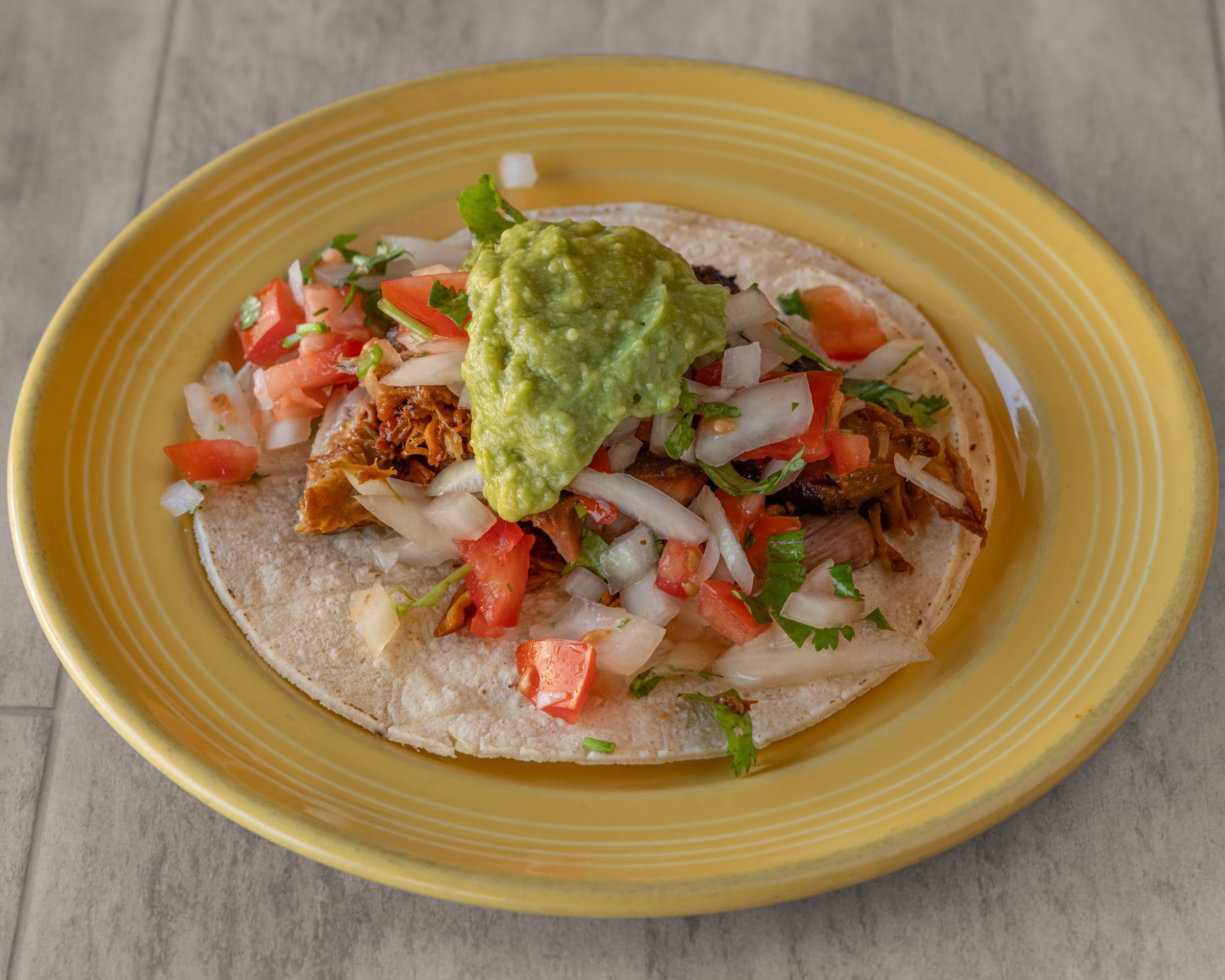 A taco with shredded meat, chopped tomato, onion, cilantro and guacamole on a yellow plate