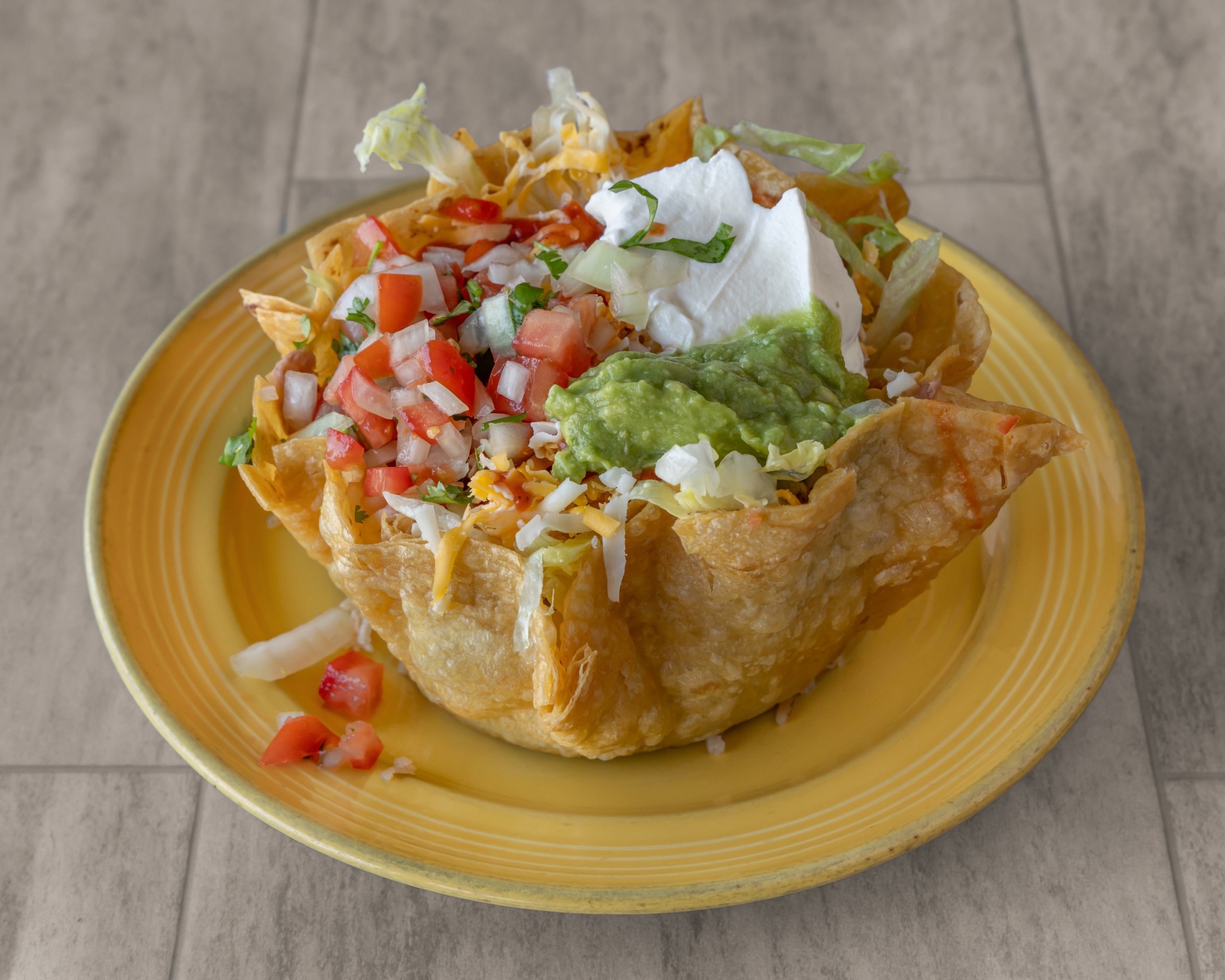 A fried tortilla bowl filled with pico de gallo, guacamole, sour cream, lettuce and cheese on a yellow plate