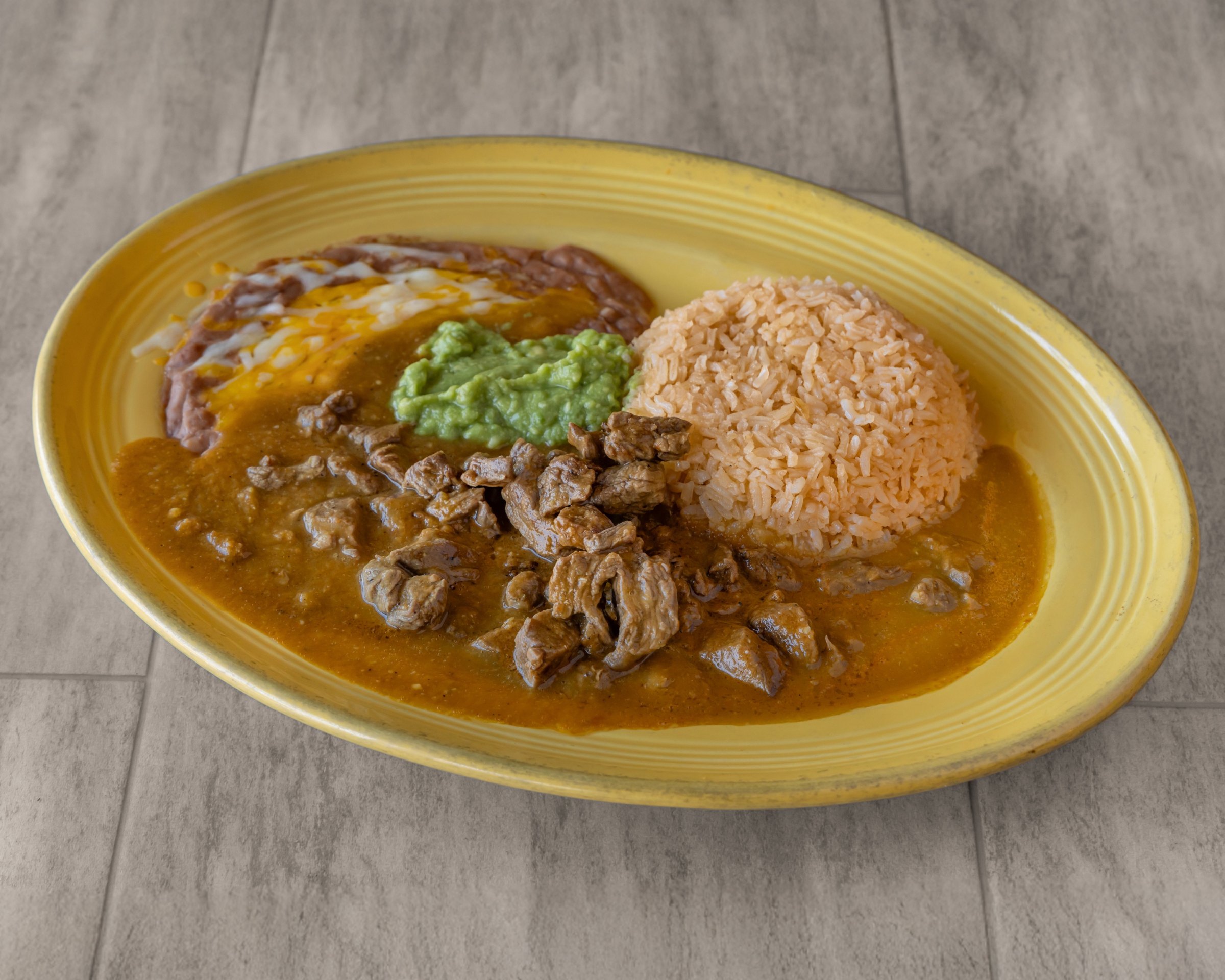 Chunks of beef in brown sauce with rice, guacamole and refried beans with cheese on a yellow plate