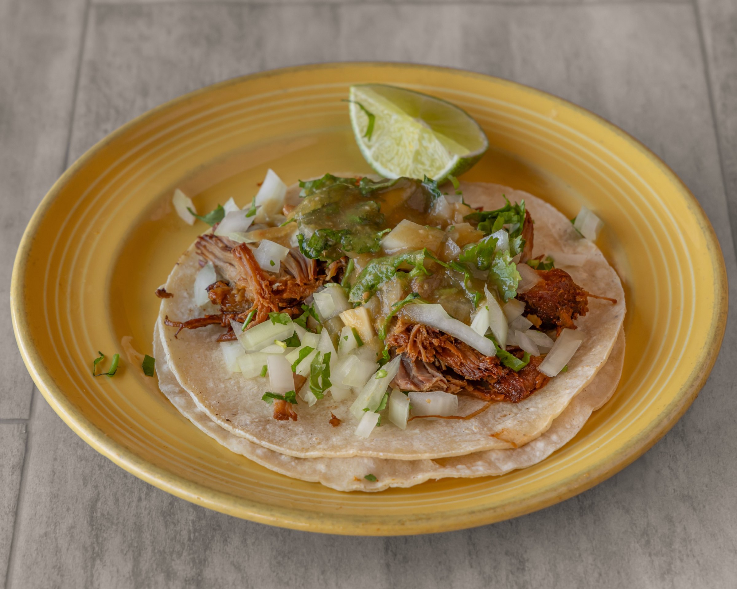 A shredded-meat taco with onion, cilantro, green salsa and a lime wedge on a yellow plate