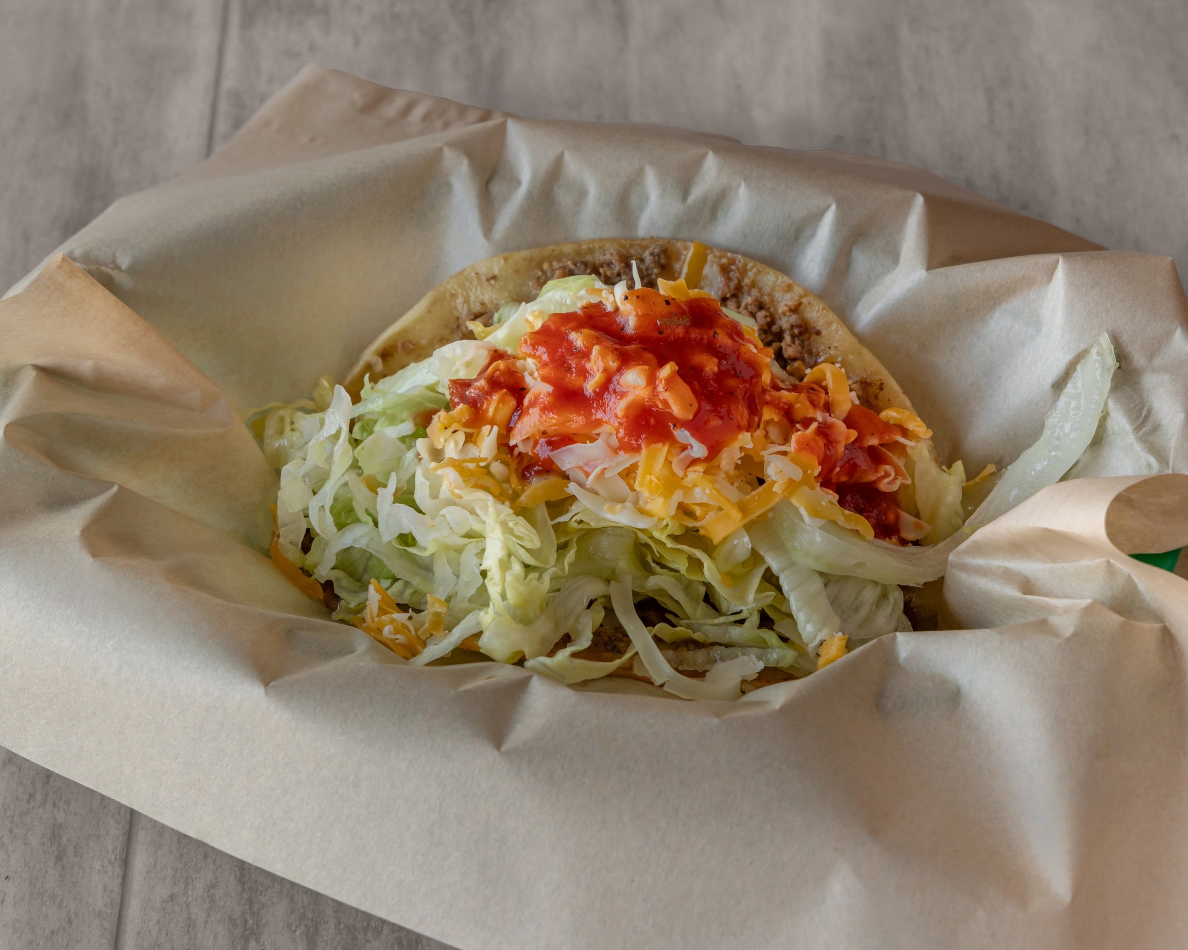 A crispy taco in paper topped with lettuce, shredded cheese and red salsa