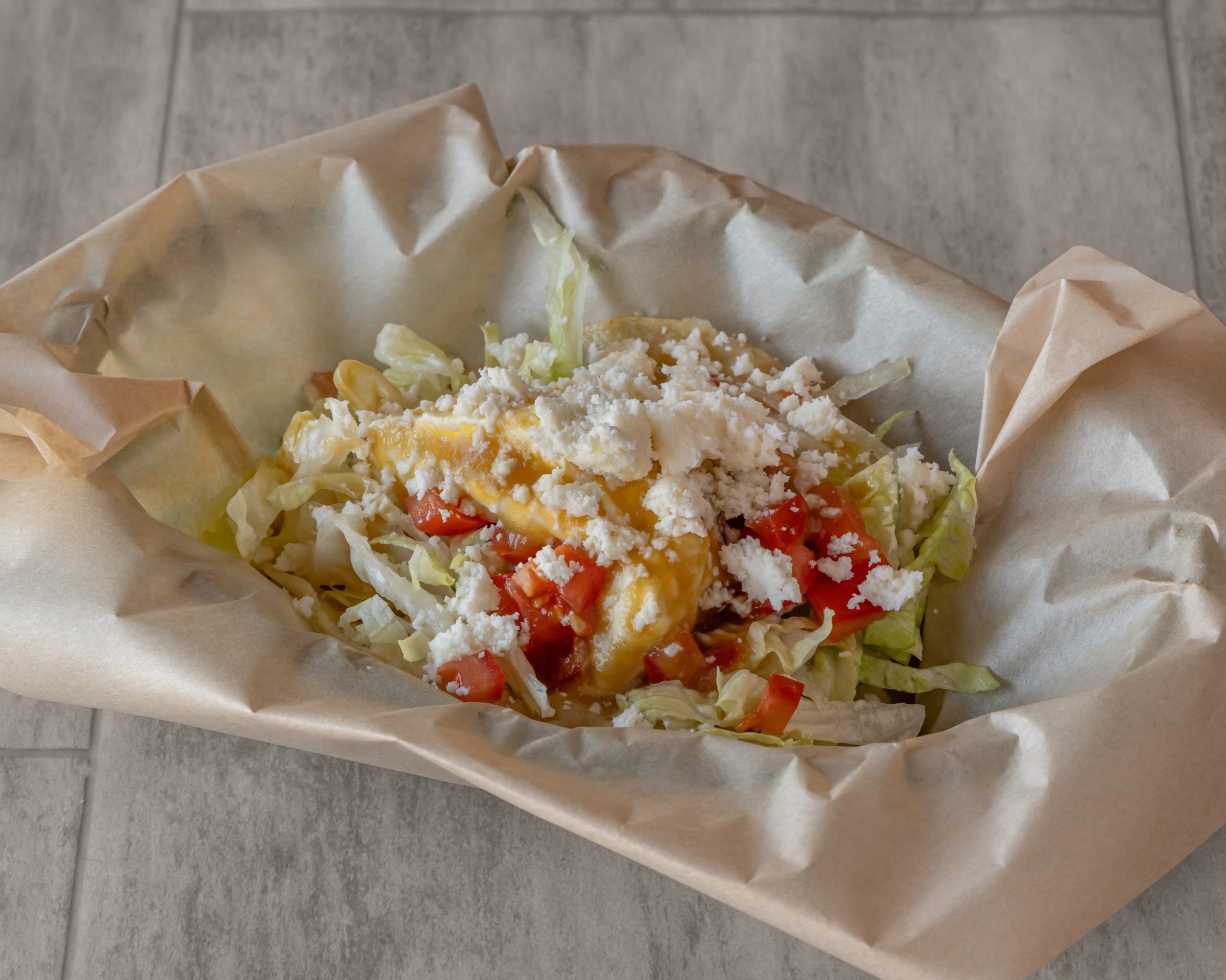 A taco in paper topped with crumbled white cheese, chopped tomato and lettuce