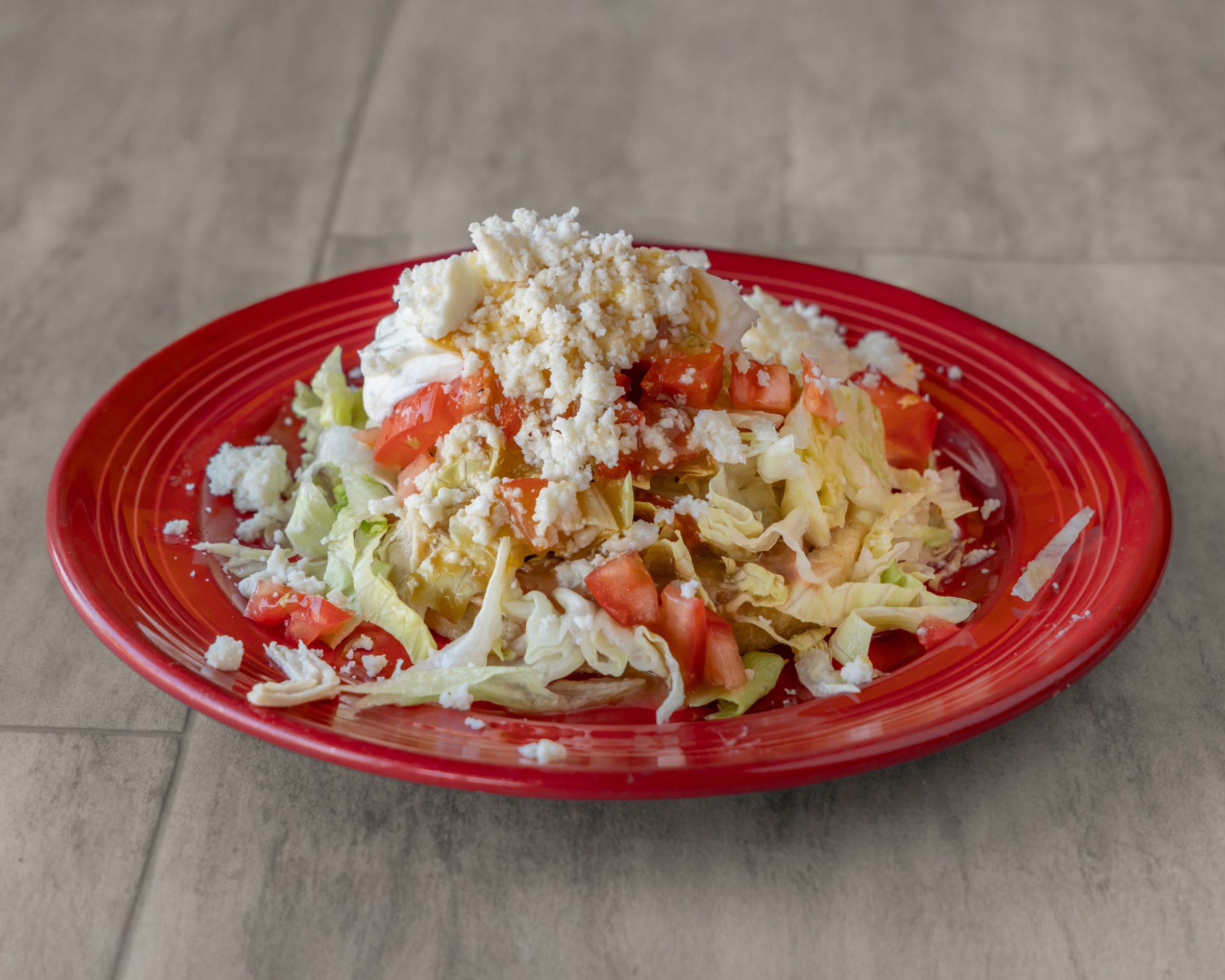 A fried tortilla topped with lettuce, chopped tomato, sour cream and crumbled white cheese on a red plate