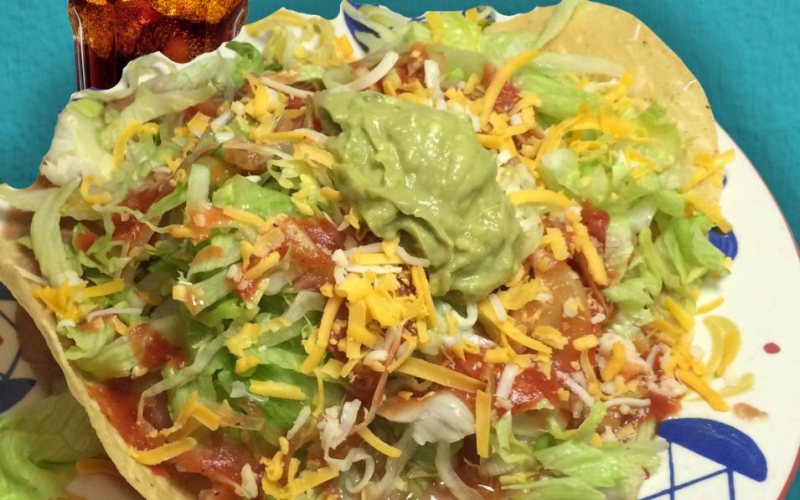 A tostada piled with lettuce, shredded cheese, red sauce and a scoop of guacamole