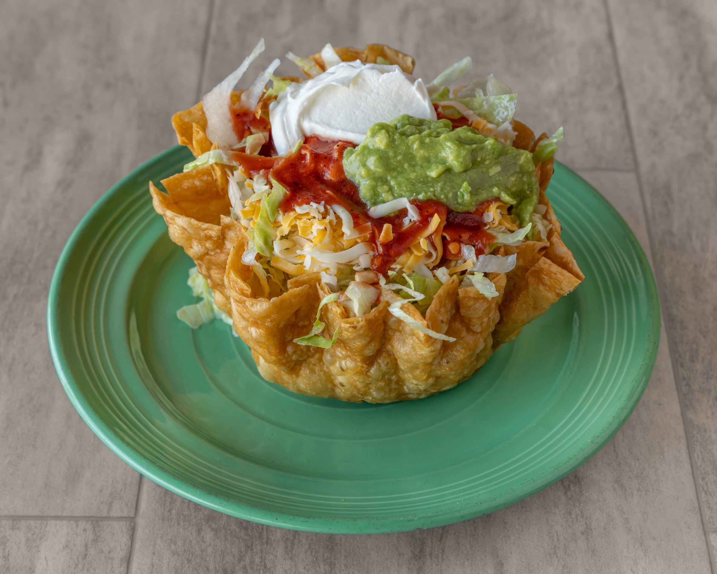 A fried tortilla bowl with lettuce, cheese, red salsa, guacamole and sour cream on a green plate